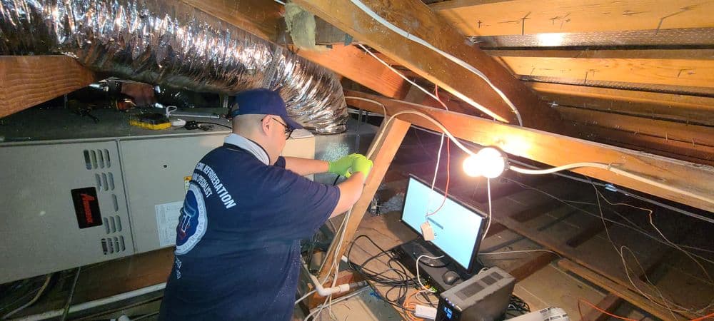 Technician inspecting HVAC system in attic with computer and equipment present.