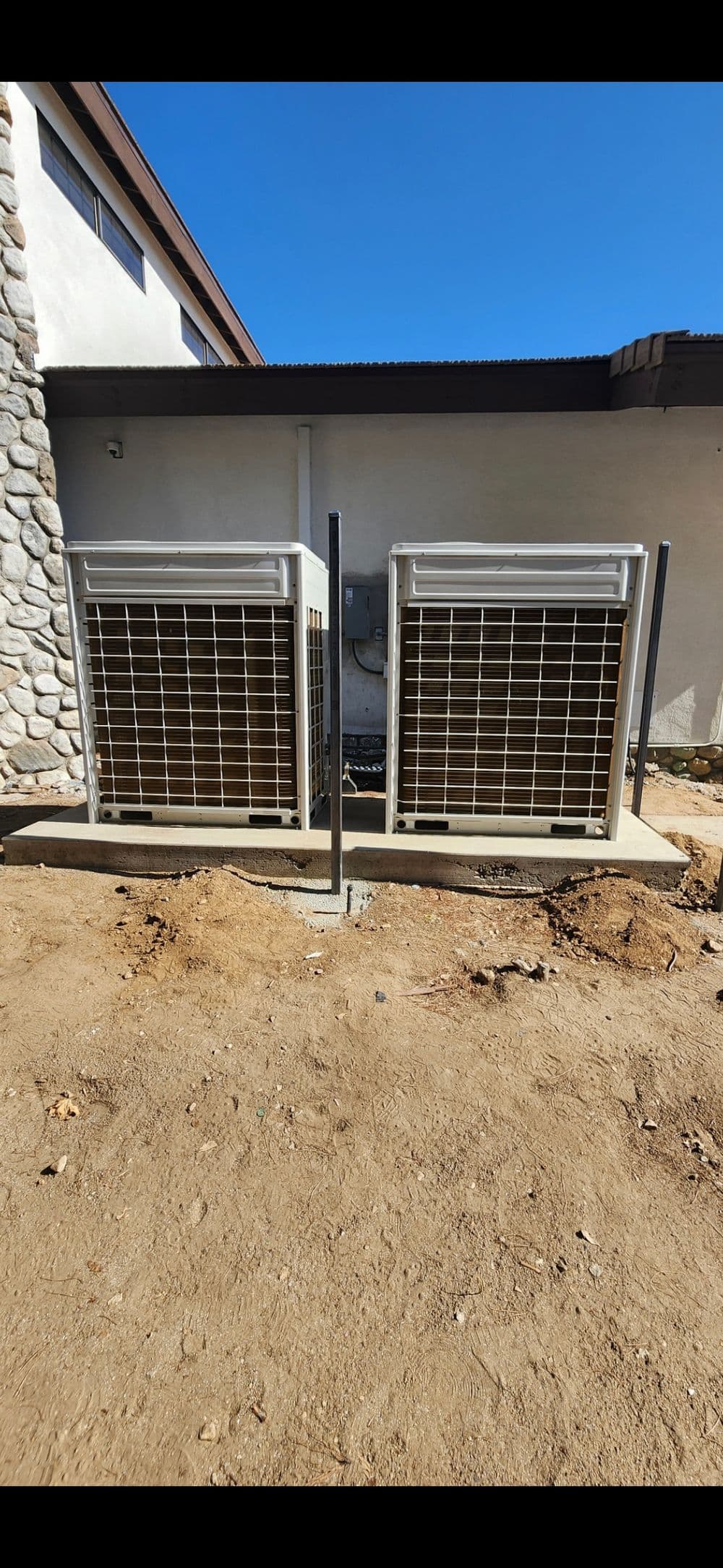 Two outdoor air conditioning units secured with metal grates, set against a stone and stucco wall.