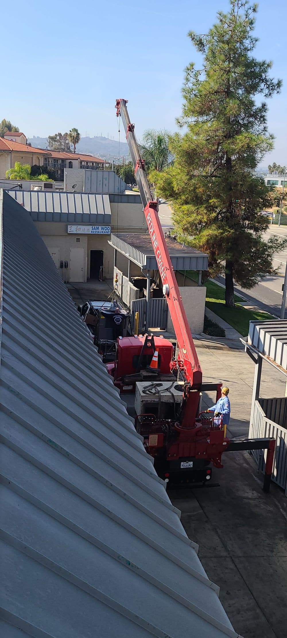 Crane lifting equipment at a commercial building, with trees and blue sky in the background.
