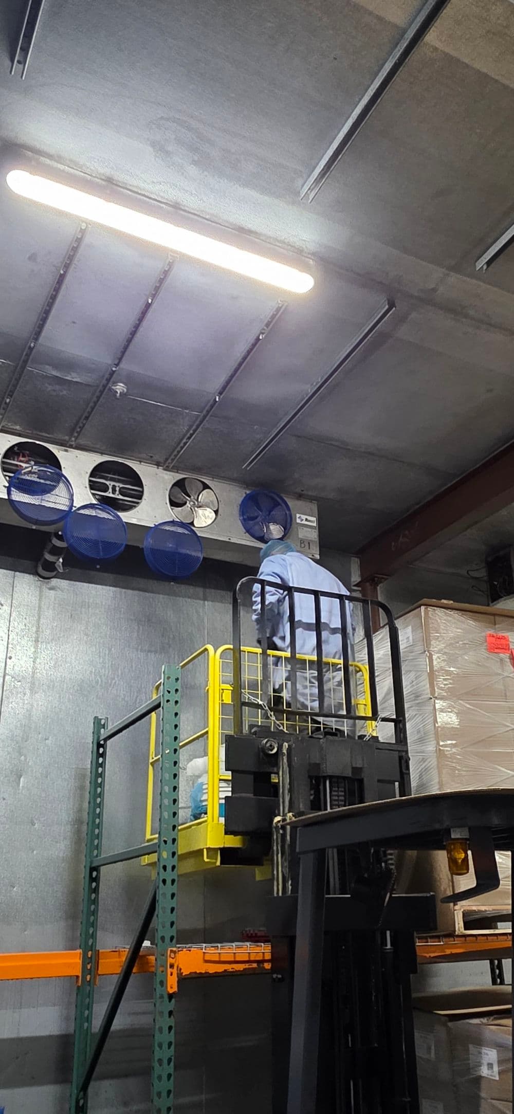Warehouse worker operating a forklift to access high shelves with ventilation system overhead.
