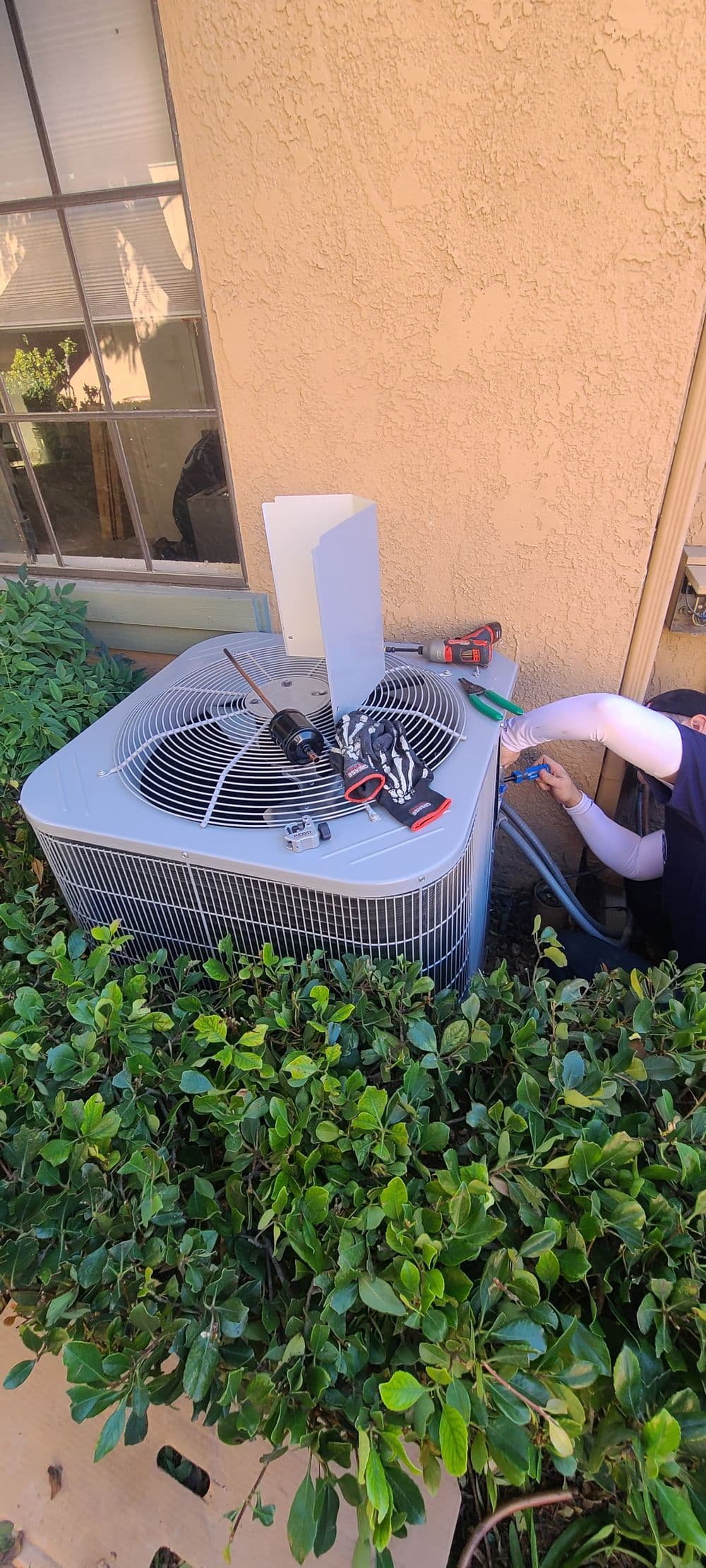 Technician servicing air conditioning unit beside a house, with tools and foliage nearby.