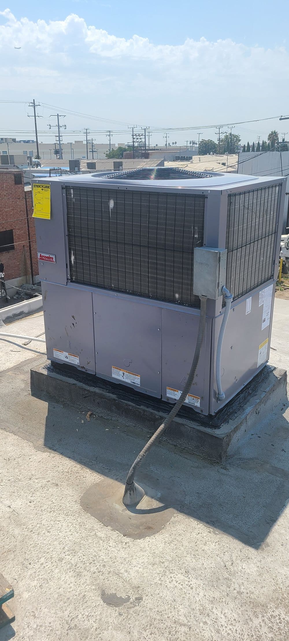 Rooftop commercial air conditioning unit with visible electrical connections and surrounding cityscape.