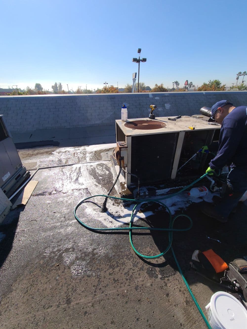 Technician cleaning rooftop air conditioning unit with hose and equipment on a sunny day.