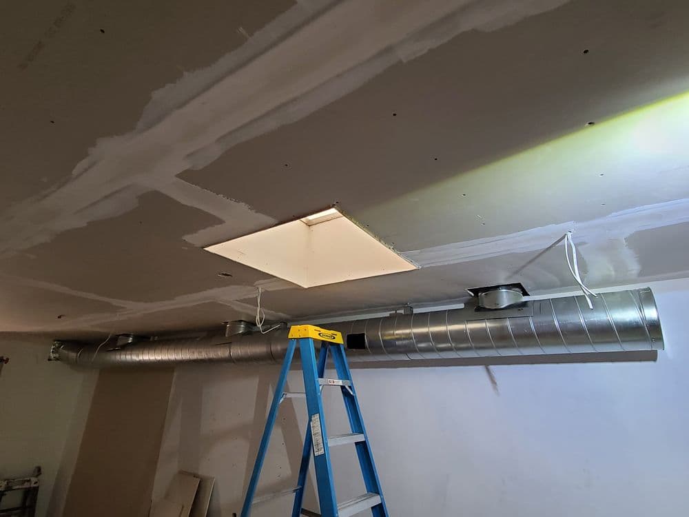 Ceiling ductwork installation with ladder, exposed drywall, and light fixture.