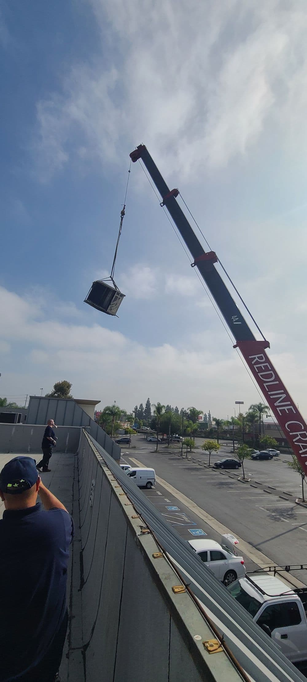 Crane lifting a large object over a parking lot with workers observing from a rooftop.