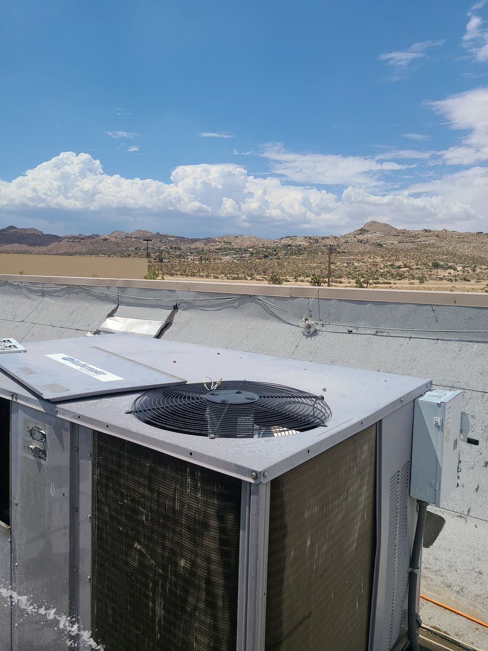 Rooftop air conditioning unit against a desert landscape with blue sky and clouds.