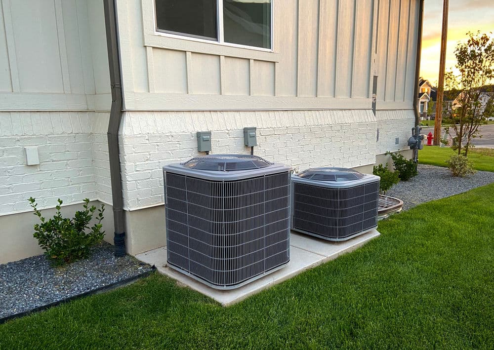 Two air conditioning units beside a house, with green grass and gravel landscaping.